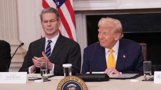 Donald Trump, seated, smiles behind a presidential seal and a tabletop microphone. He is wearing a navy suit jacket and gold-colored tie. To his right sits Treasury Secretary Scott Bessent, who is clapping and wearing a dark suit jacket and blue striped tie. Behind them a US flag stands.
