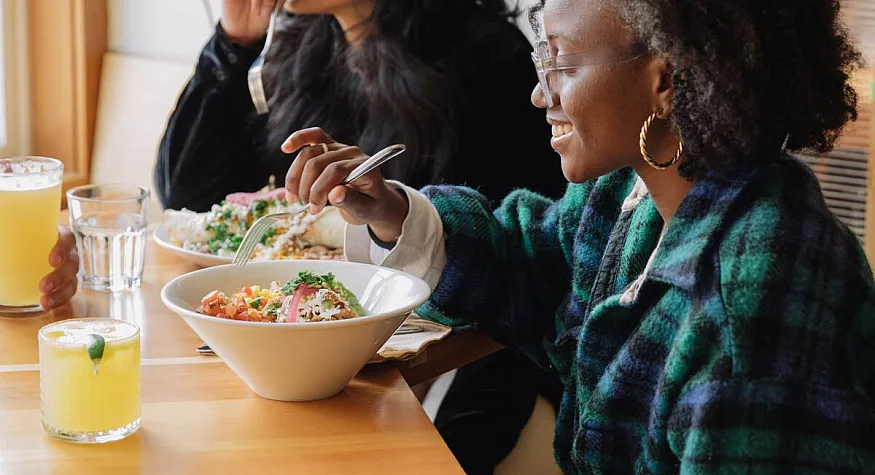 Two people, one in a wheelchair, sit at a wooden table eating bowls of food with drinks on the table.