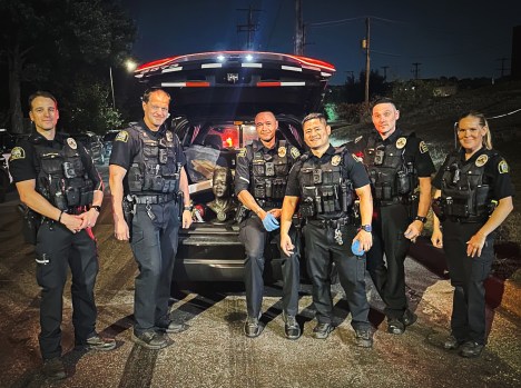 STP-L-SUNI-0829 Six uniformed officers pose around a Suni Lee bust in the back of a police SUV.